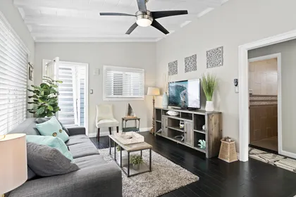 Vantage point from kitchen toward living and entry areas with TV, ceiling fan and comfy seating. Hardwood flooring throughout.