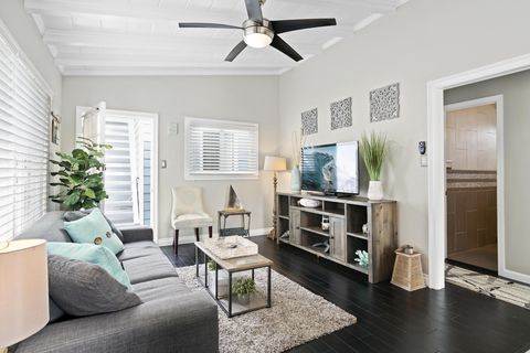 Vantage point from kitchen toward living and entry areas with TV, ceiling fan and comfy seating. Hardwood flooring throughout.